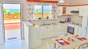 A kitchen with a view of the sea, white cabinets, a dining table with chairs, and a hanging light fixture. - Eliana (Kissamos)