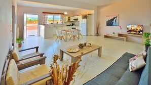 A spacious living area with a sofa, a coffee table, and a dining table. There is a kitchen in the background with a view of the outdoors. - Eliana (Kissamos)