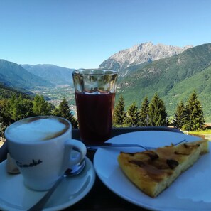 A table with a cup of coffee, a glass of red beverage, and a slice of pizza, set against a mountainous backdrop. - Albergo Sargas (Cevo)