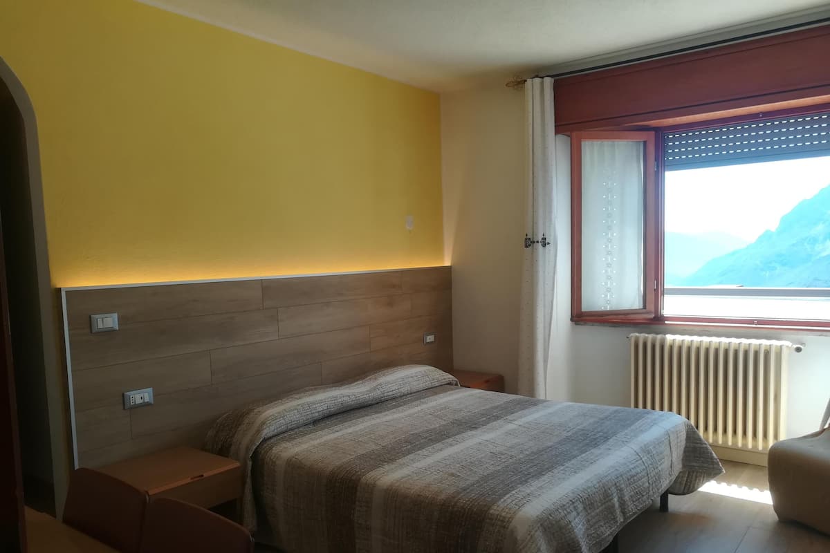 A bedroom with a wooden headboard, a bed with a striped blanket, a bedside table, a nightstand, a window with a view of mountains, and a radiator.