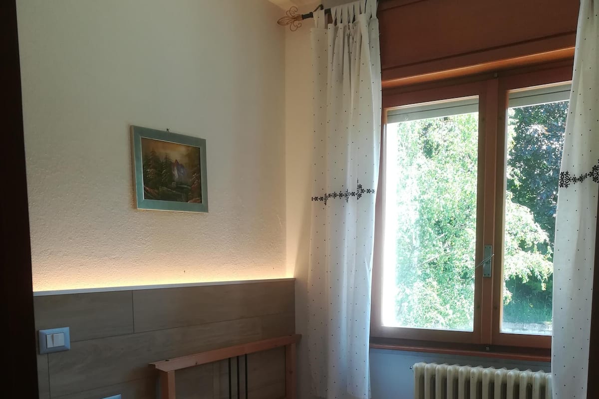A bedroom with a bed, a wooden dresser, a radiator, a window with curtains, and a painting on the wall.
