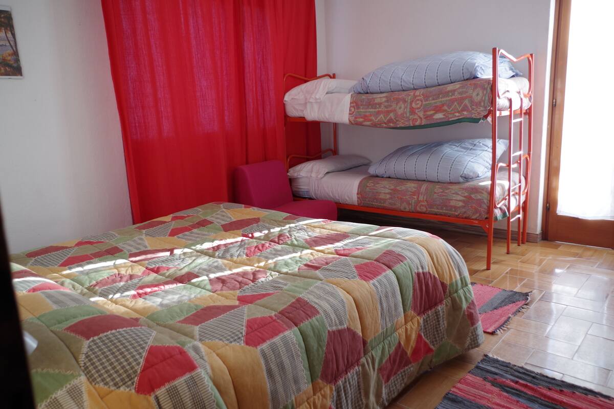 A room with a bunk bed, a single bed with a colorful quilt, a red curtain, and a wooden floor.