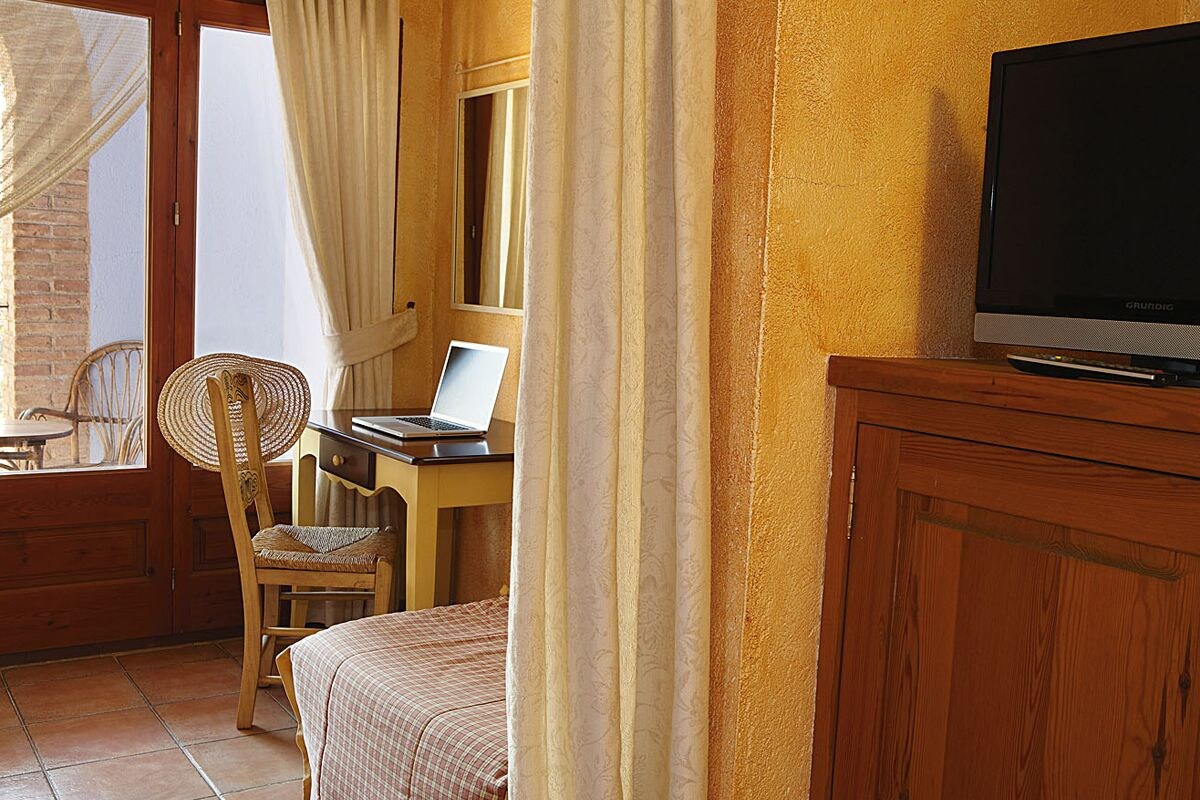 Ein Hotelzimmer mit einem Holztisch, einem Laptop, einem Korbstuhl, einem Bett und einem Fernseher auf einer Holzkommode.