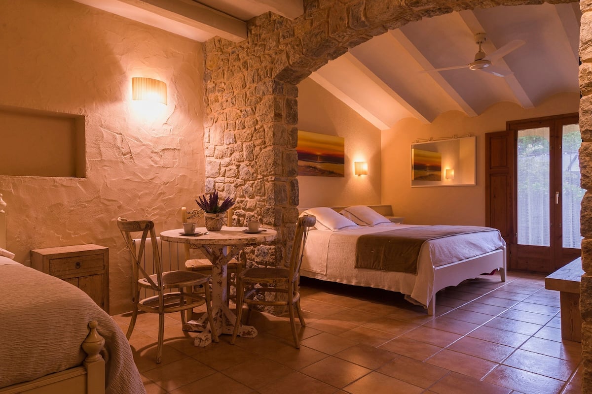 Ein gemütliches Schlafzimmer mit einem Steinbogen, einem Bett, einem Nachttisch mit Lampe und Blick ins Freie durch ein Fenster.