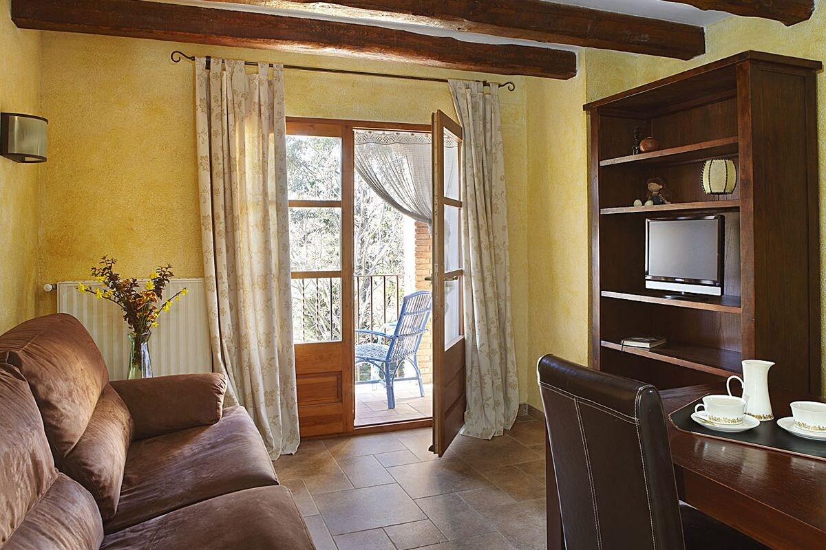 Ein gemütliches Zimmer mit einem braunen Sofa, einem Esstisch mit einer Karaffe und Tassen, einem Fernseher auf einem Holzregal und einer Tür, die auf einen Balkon mit einem Stuhl führt.
