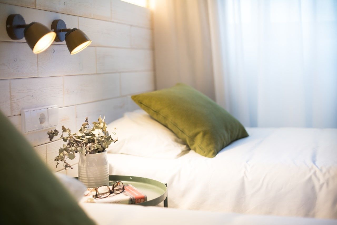 Una cama bien hecha con ropa blanca y una almohada verde, una mesita de noche con un jarrón de flores secas y dos lámparas fijadas a la pared.