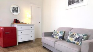Una habitación con un sofá gris, dos almohadas estampadas, un refrigerador rojo de estilo vintage, un tocador blanco con cajones, un espejo redondo y un cuadro en la pared.