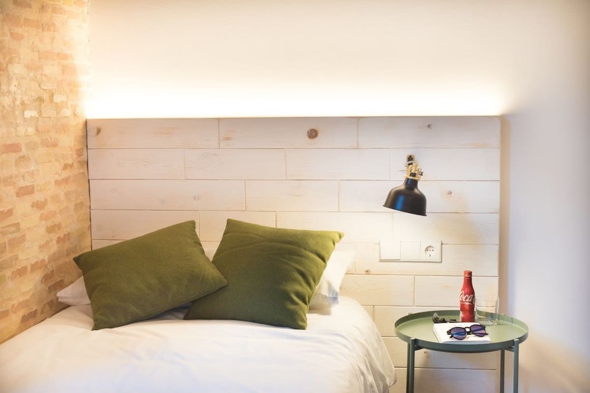 A bedroom with a white bed, green pillows, a small round table with a bottle and glasses, and a brick wall.