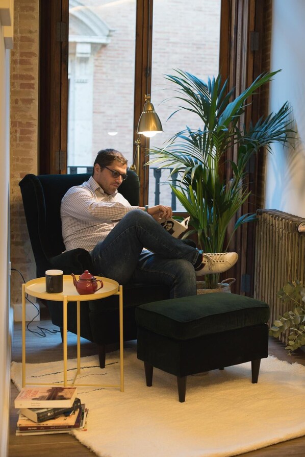 Un hombre sentado en un sillón oscuro junto a una ventana, con una planta a su lado. Hay una mesita con un hervidor y una taza, y un radiador al fondo.