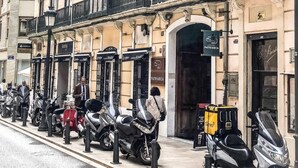 Una fila de scooters y motocicletas estacionadas a lo largo de una acera frente a un edificio con detalles arquitectónicos ornamentados y varios balcones.