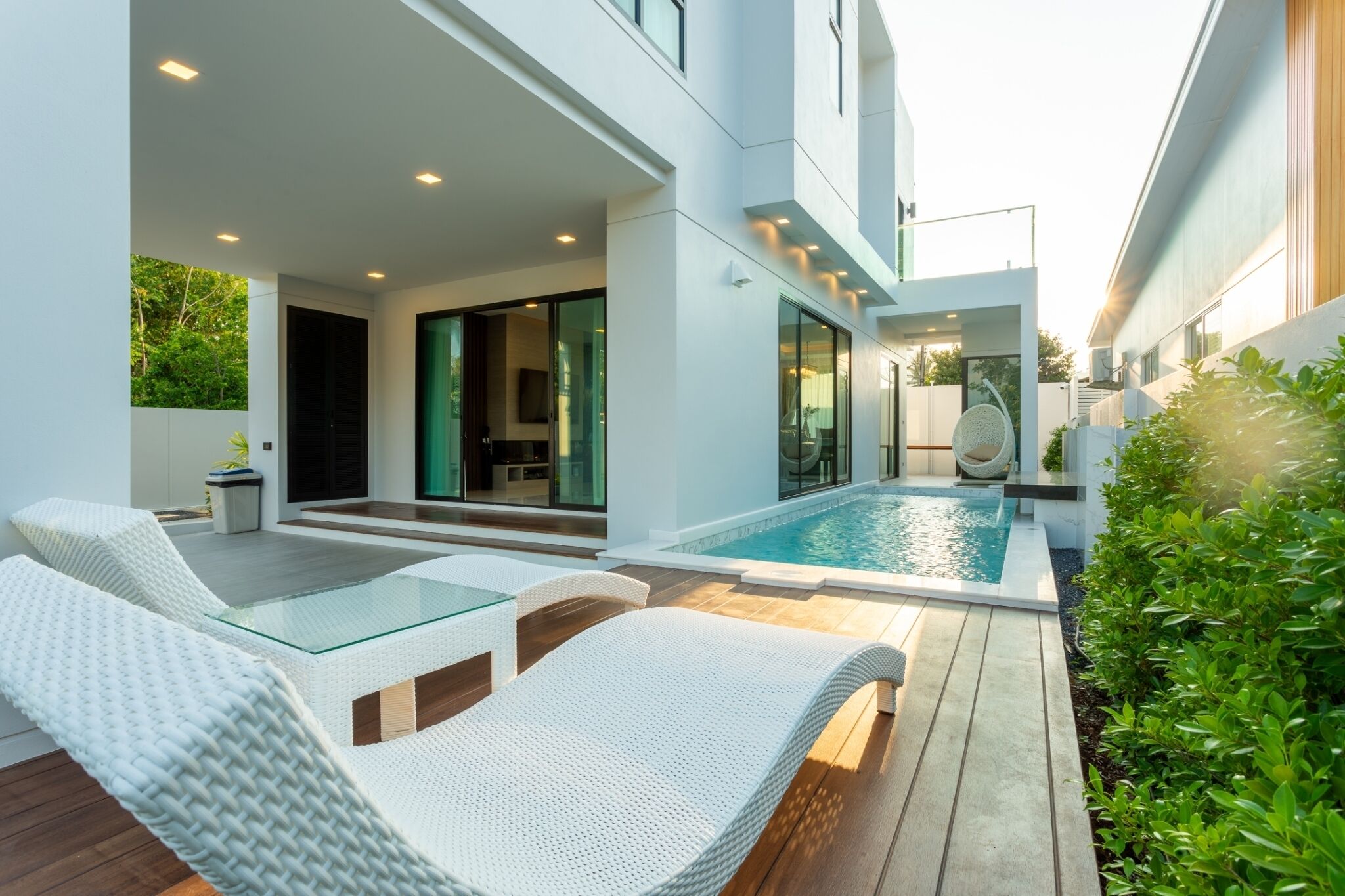 A modern outdoor pool area with white wicker lounge chairs, a glass table, and a clear view of the pool.