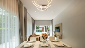 A modern dining area with a long table set for four, a sofa in the background, and a view of a pool and greenery outside. - The Haven Krabi (Krabi)
