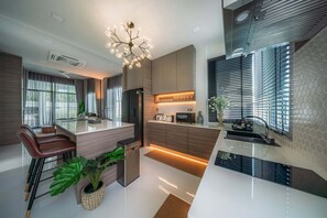 A modern kitchen with a central island, a dining area with a brown leather chair, and a large window with blinds. - The Haven Krabi (Krabi)