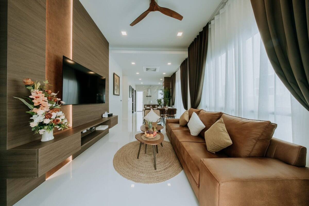 A modern living room with a brown leather sofa, a flat-screen TV mounted on a wooden wall, a dining area in the background, and a ceiling fan.