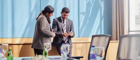 Two people in business attire standing in a conference room with a table set for a meeting, featuring a laptop, notebooks, and a water bottle.