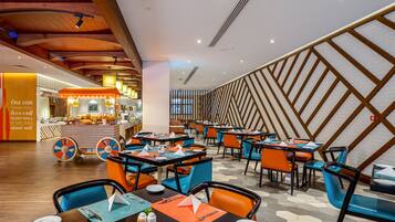 A modern restaurant with a central bar, multiple dining tables with orange and blue chairs, and a geometric patterned wall.