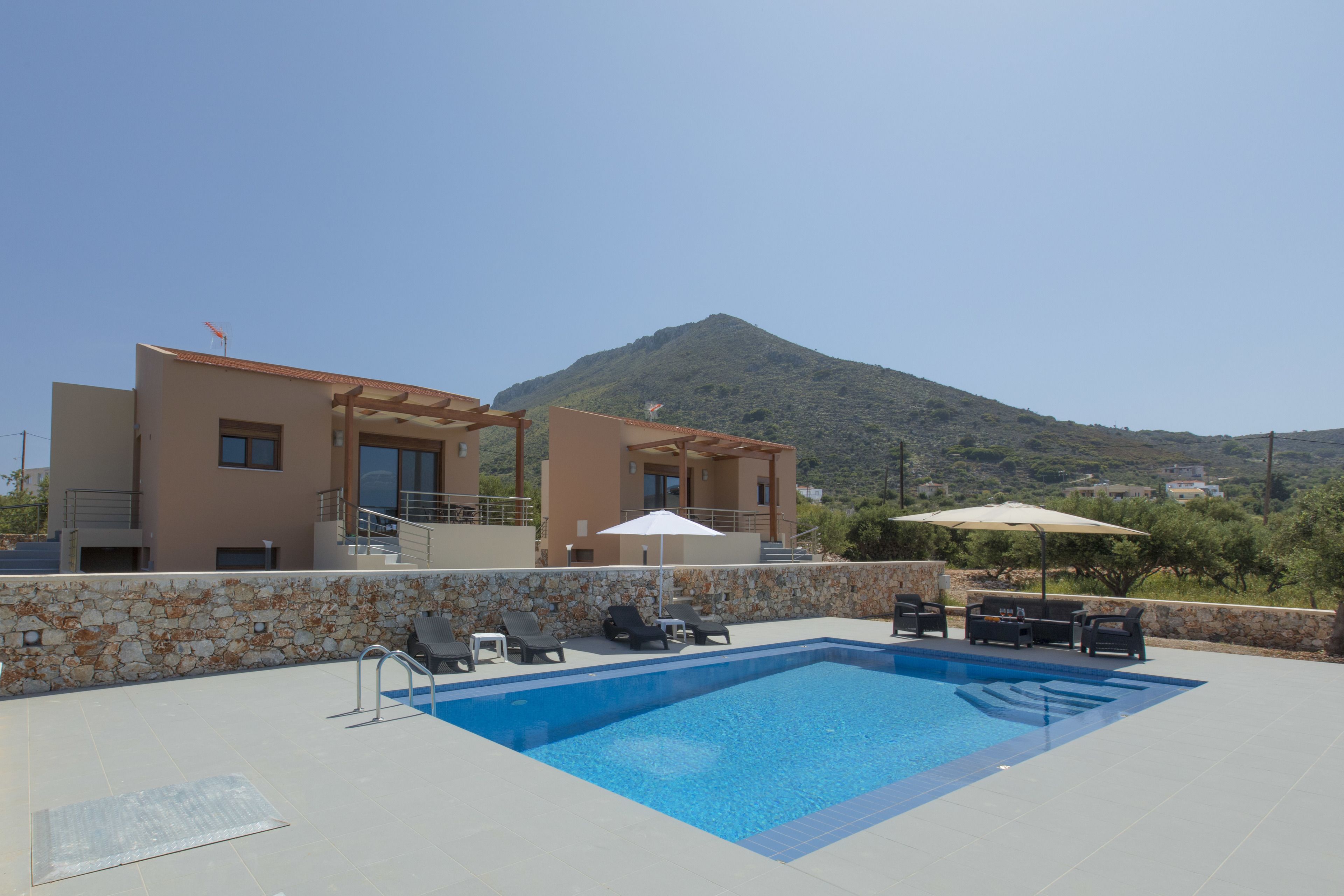 A modern poolside area with lounge chairs and umbrellas, a stone wall, and a mountain in the background.