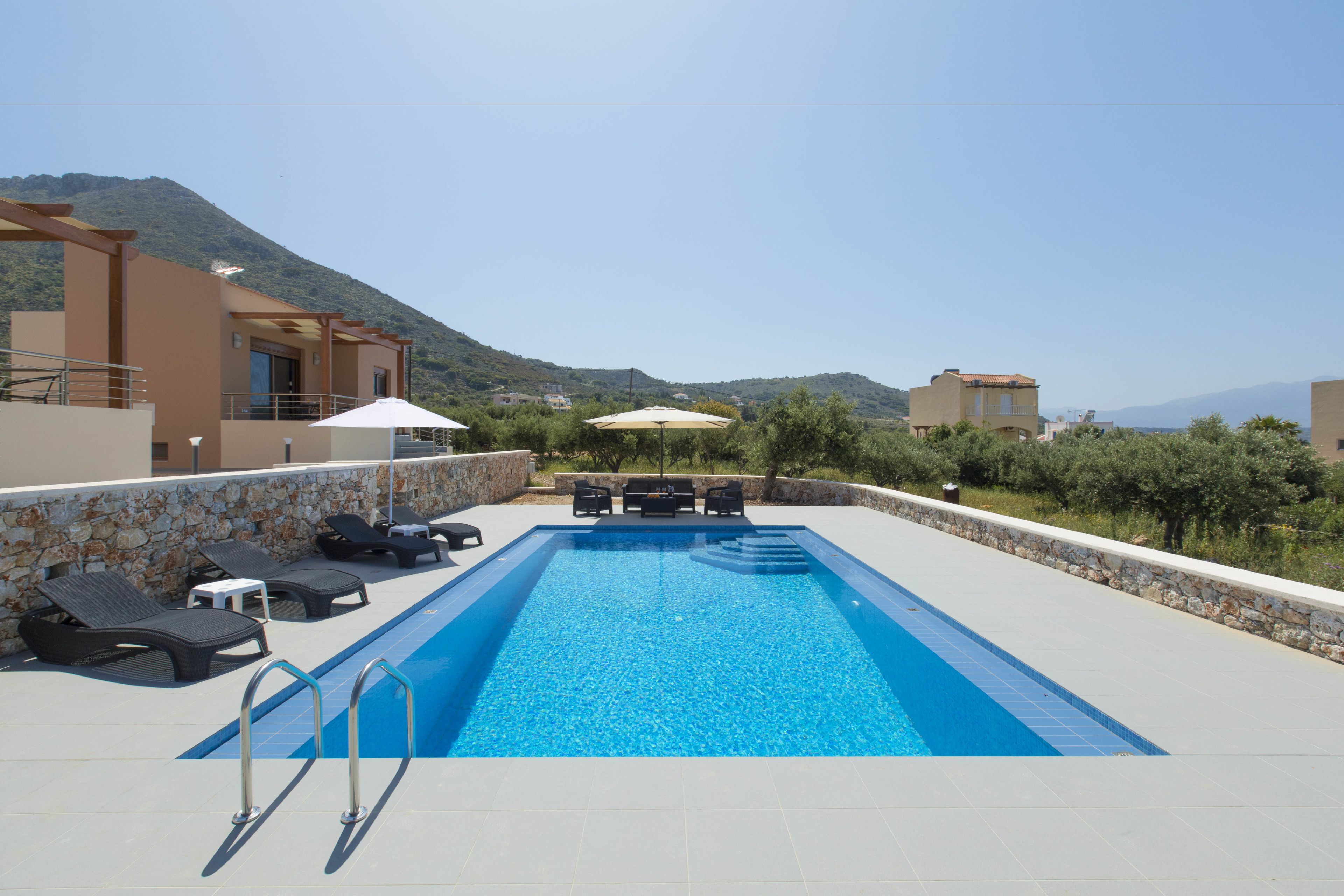 Una piscina con sillas de descanso y sombrillas, rodeada de muros de piedra y vista a la montaña.