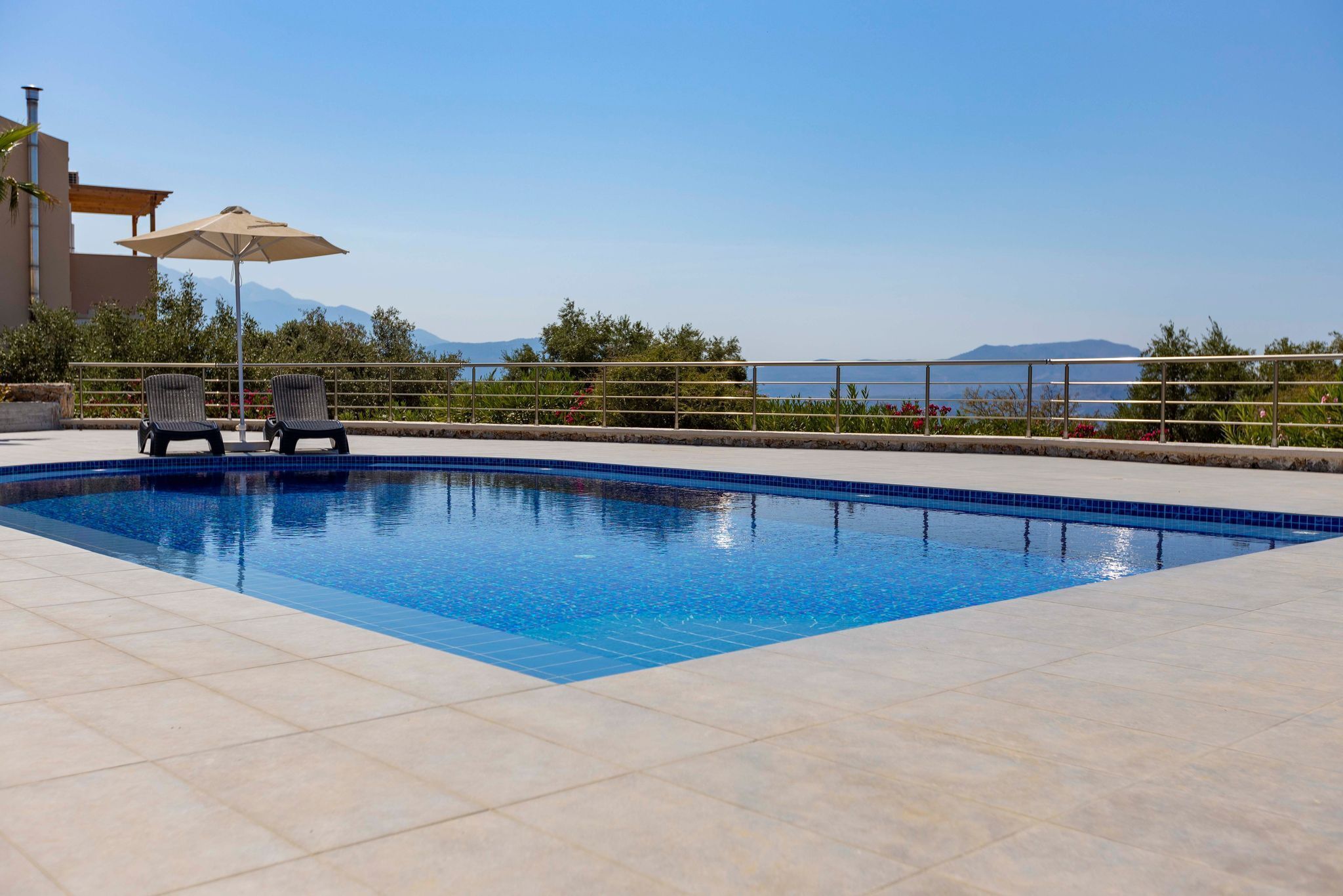 A swimming pool with two lounge chairs and an umbrella, surrounded by a tiled deck and a railing, with a view of mountains and the sea in the background.
