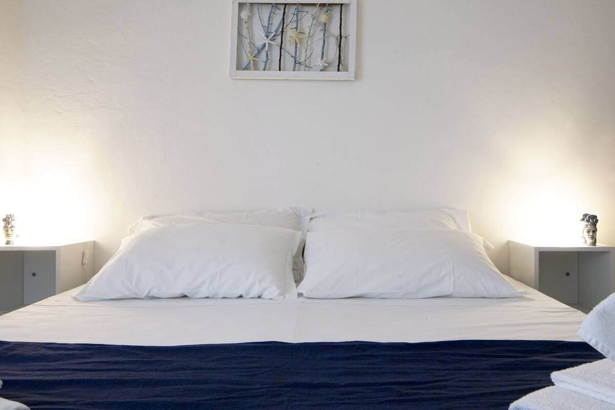 A neatly made bed with white linens, two bedside tables with decorative items, and a framed artwork above the bed.