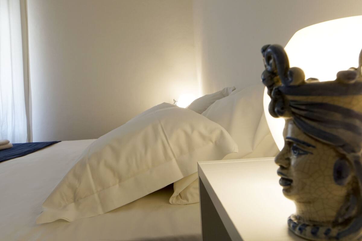 A hotel room with a neatly made bed, a statue on the nightstand, and a window with curtains.