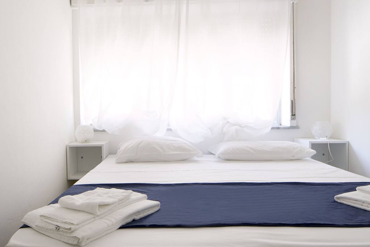 A neatly made bed with white pillows and a blue blanket, a bedside table with a lamp, and sheer curtains on a window.