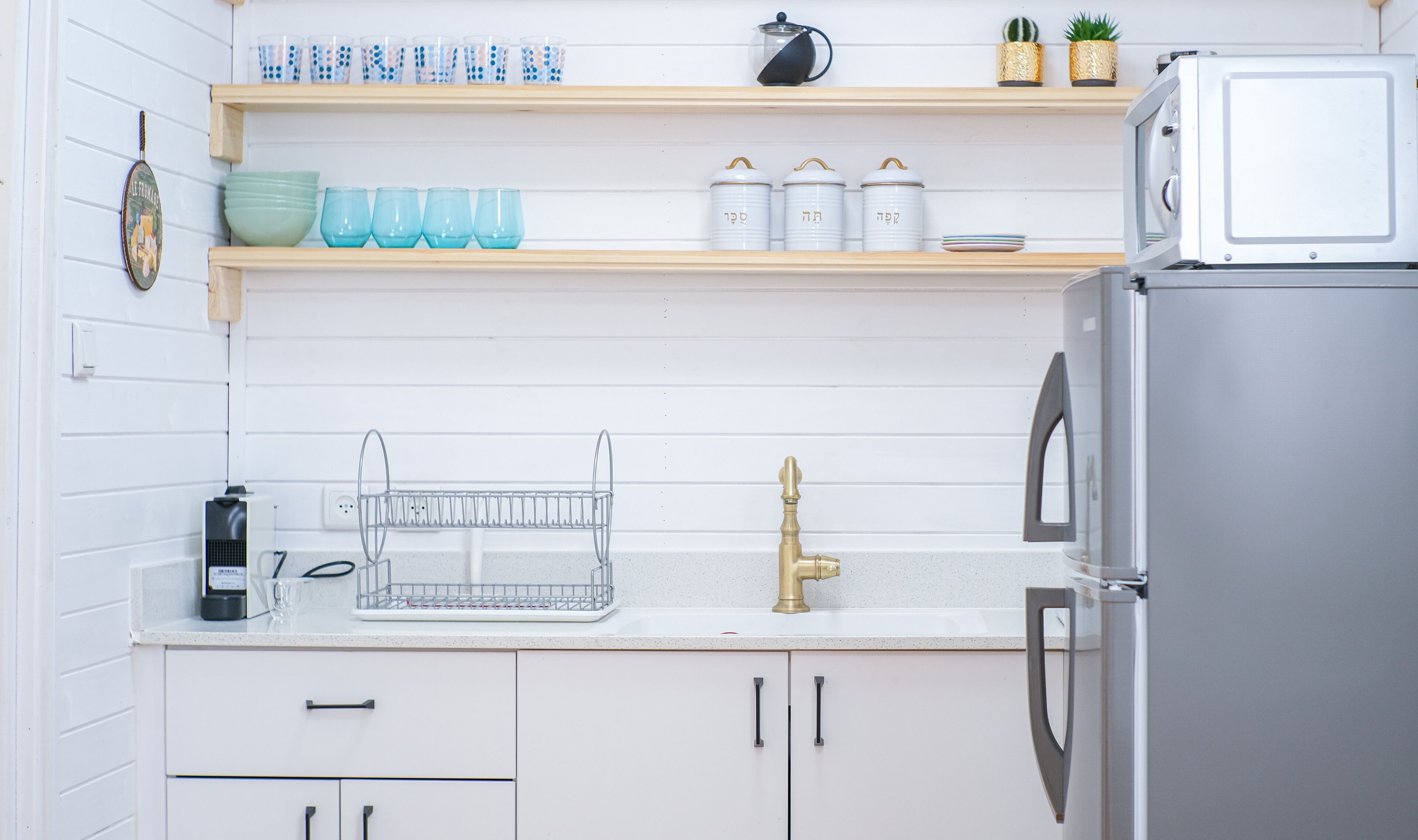 A kitchen with white cabinets, a stainless steel refrigerator, a sink with a gold faucet, and various kitchen items on shelves.