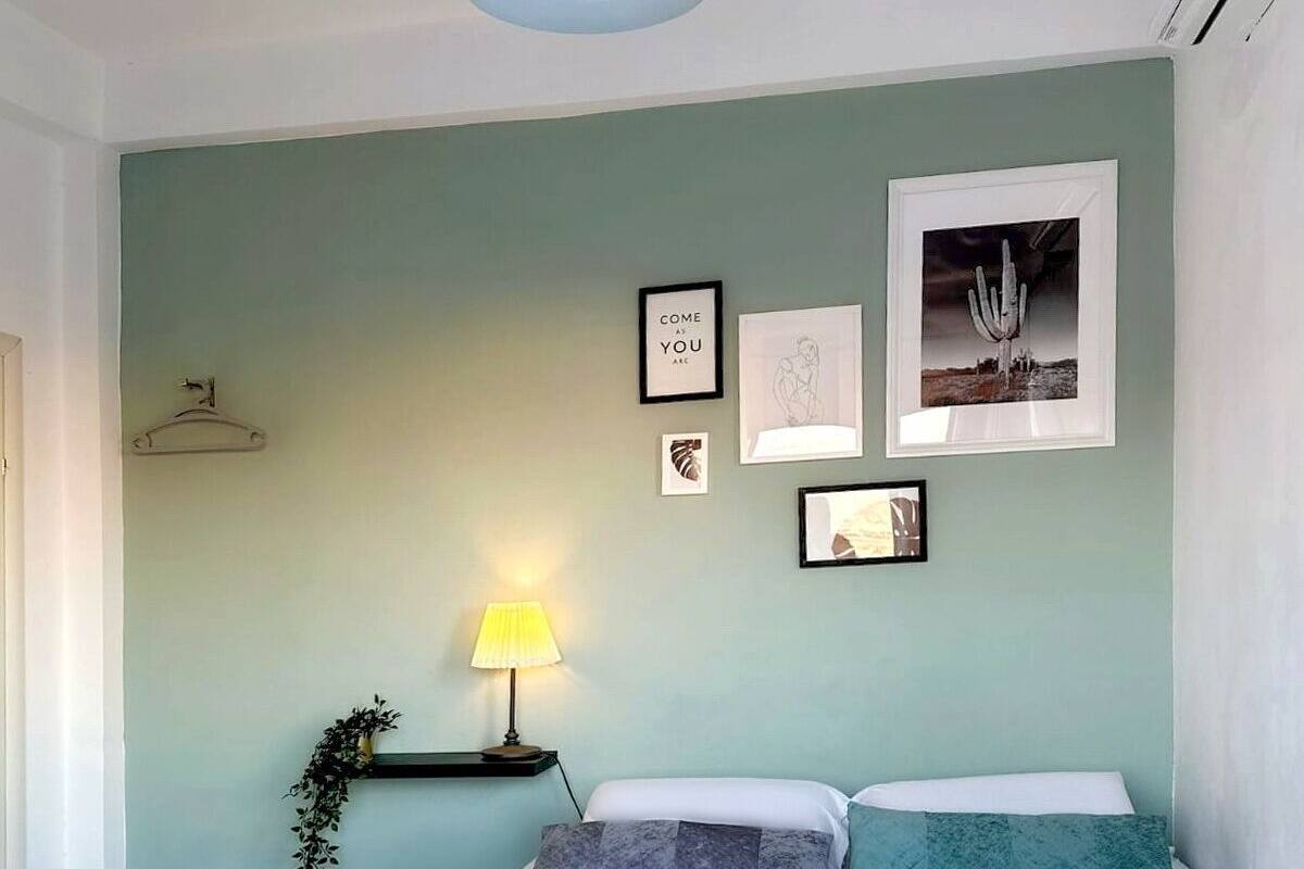A neatly made bed with rolled towels, a wall-mounted lamp, and framed artwork on a light teal wall.