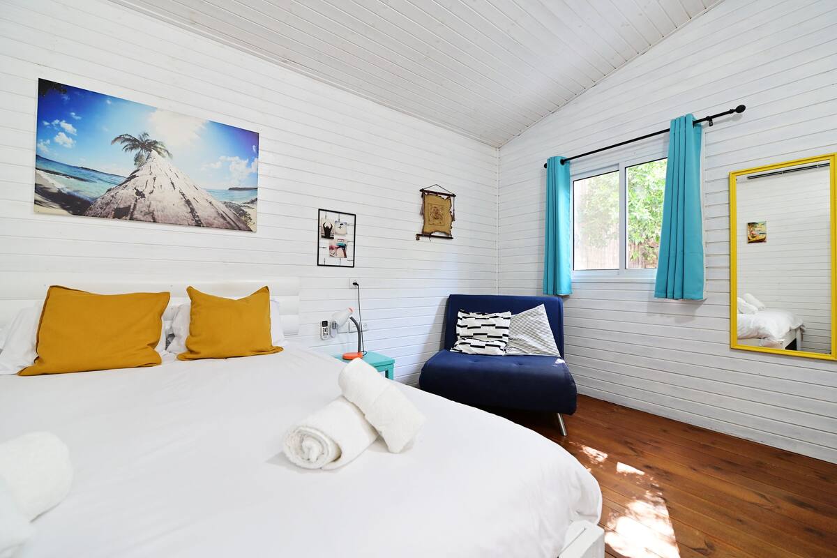 A hotel room with a bed, a blue armchair, a window with blue curtains, and a painting of a beach scene.