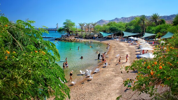 Ein Strand mit klarem türkisblauem Wasser, Menschen beim Schwimmen und Sonnenbaden, und eine Brücke im Hintergrund.