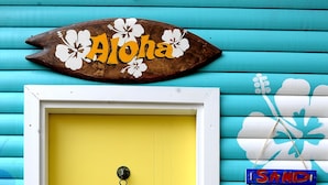Eine gelbe Tür mit weißem Rahmen, ein Holzschild darüber mit der Aufschrift "Aloha", und eine Wand mit floralem Muster sowie Schildern, die "SAND", "STRAND", "ENTSPANNEN" und "SURFEN" zeigen.