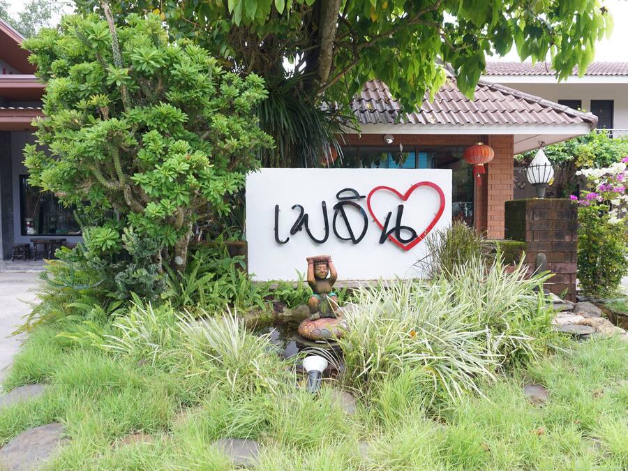 Une enseigne avec des caractères thaïlandais et un symbole en forme de cœur, entourée d’une végétation luxuriante et avec un bâtiment traditionnel en arrière-plan.