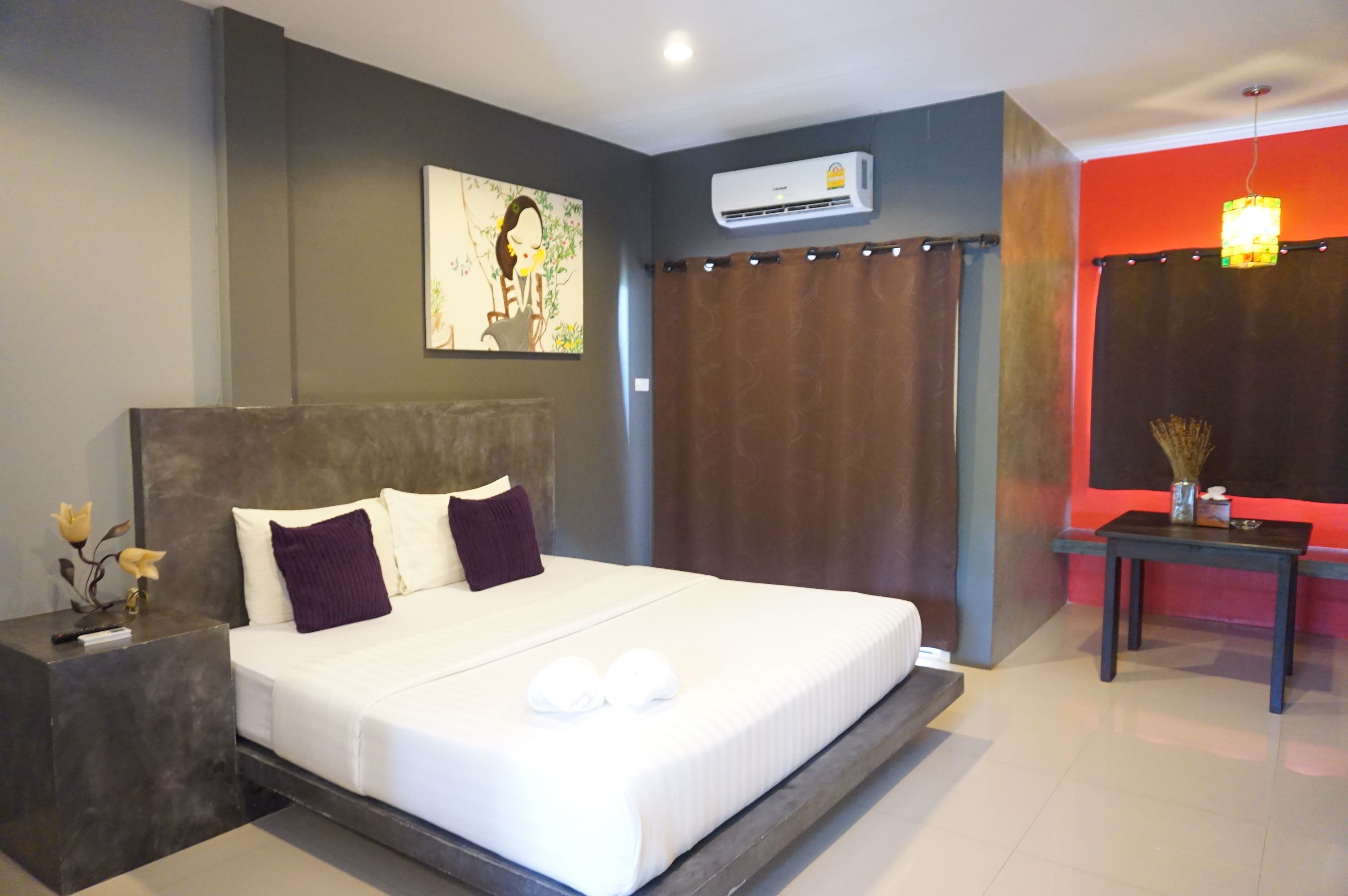 A modern hotel room with a large bed, a nightstand, an air conditioning unit, a small table with a vase, and a painting on the wall.