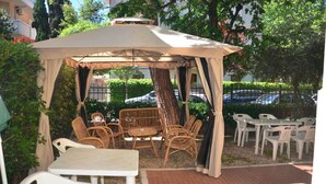 A gazebo with wicker furniture and plastic chairs, surrounded by greenery and a paved area. - Hotel Villa Pozzi (Cattolica)