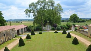 A large, open grassy area with neatly trimmed topiary trees, a central tree, and a building with a red-tiled roof. - Château des Noces (Bazoges-en-Pareds)