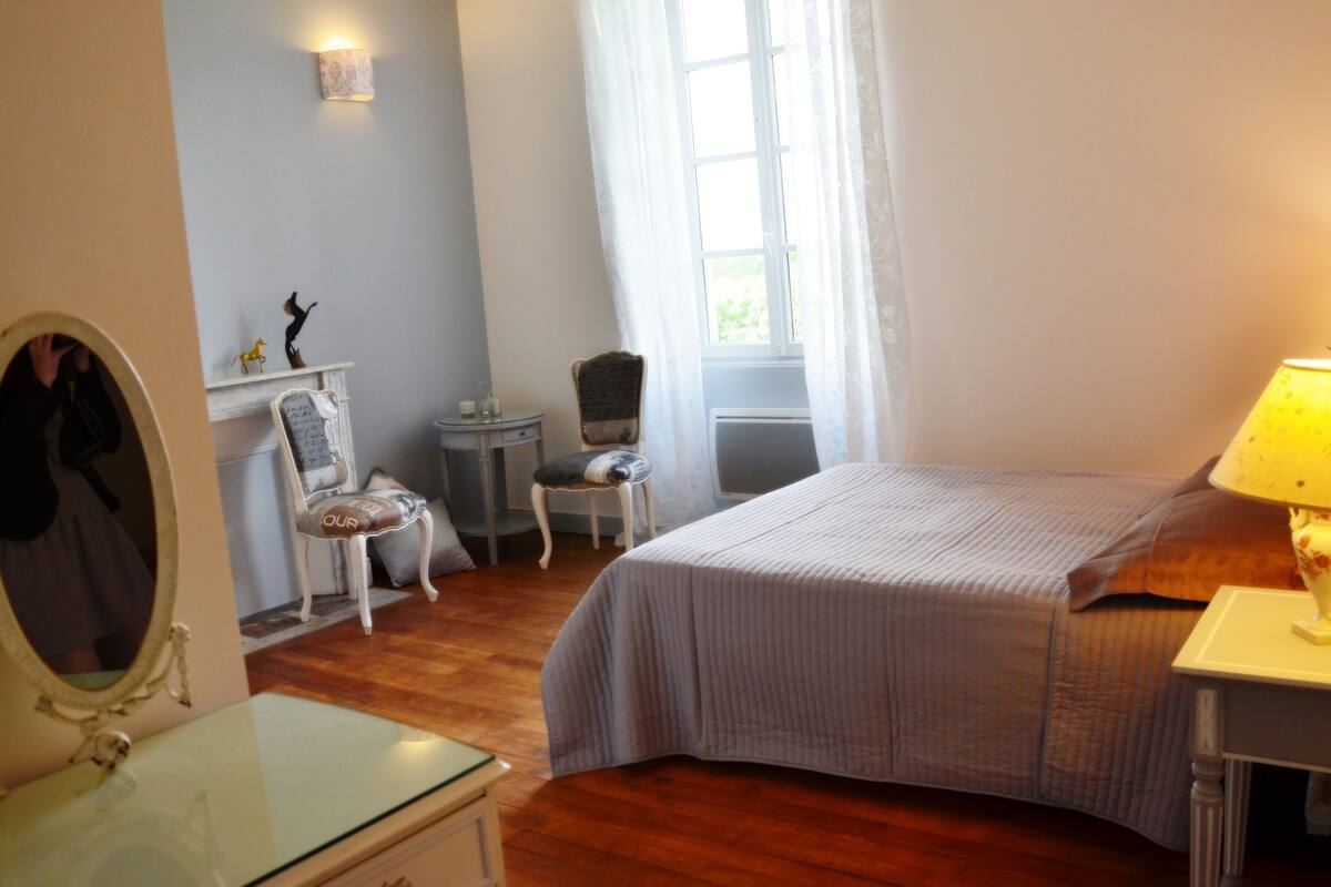 A bedroom with a bed, a dresser, a chair, a small table, a mirror, and a window with curtains.