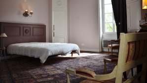 A bedroom with a bed, a chair, a nightstand, a lamp, a window with curtains, and a door with a wreath. - Château des Noces (Bazoges-en-Pareds)