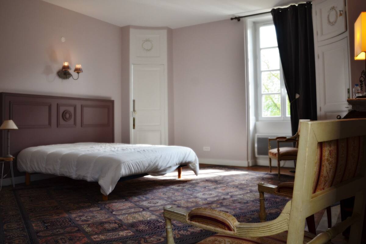 A bedroom with a bed, a chair, a nightstand, a lamp, a window with curtains, and a door with a wreath.
