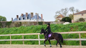 A person riding a horse in an outdoor arena with a sandy floor, surrounded by a wooden fence. In the background, there is a building with multiple chimneys and a stone wall. - Château des Noces (Bazoges-en-Pareds)