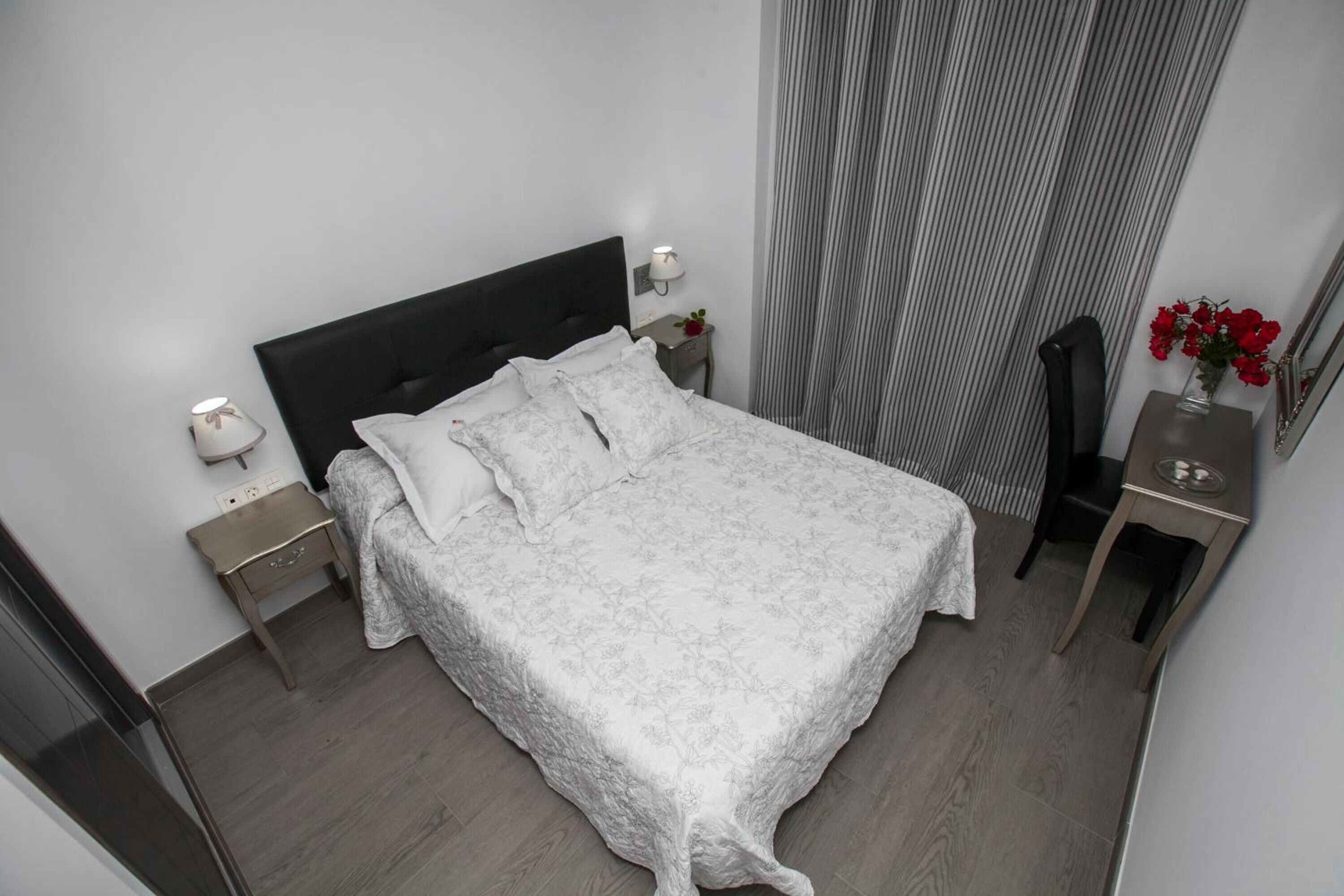 A neatly made bed with a white floral bedspread and pillows, a bedside table with a lamp, a small side table with a vase of red flowers, and a mirror on the right side.