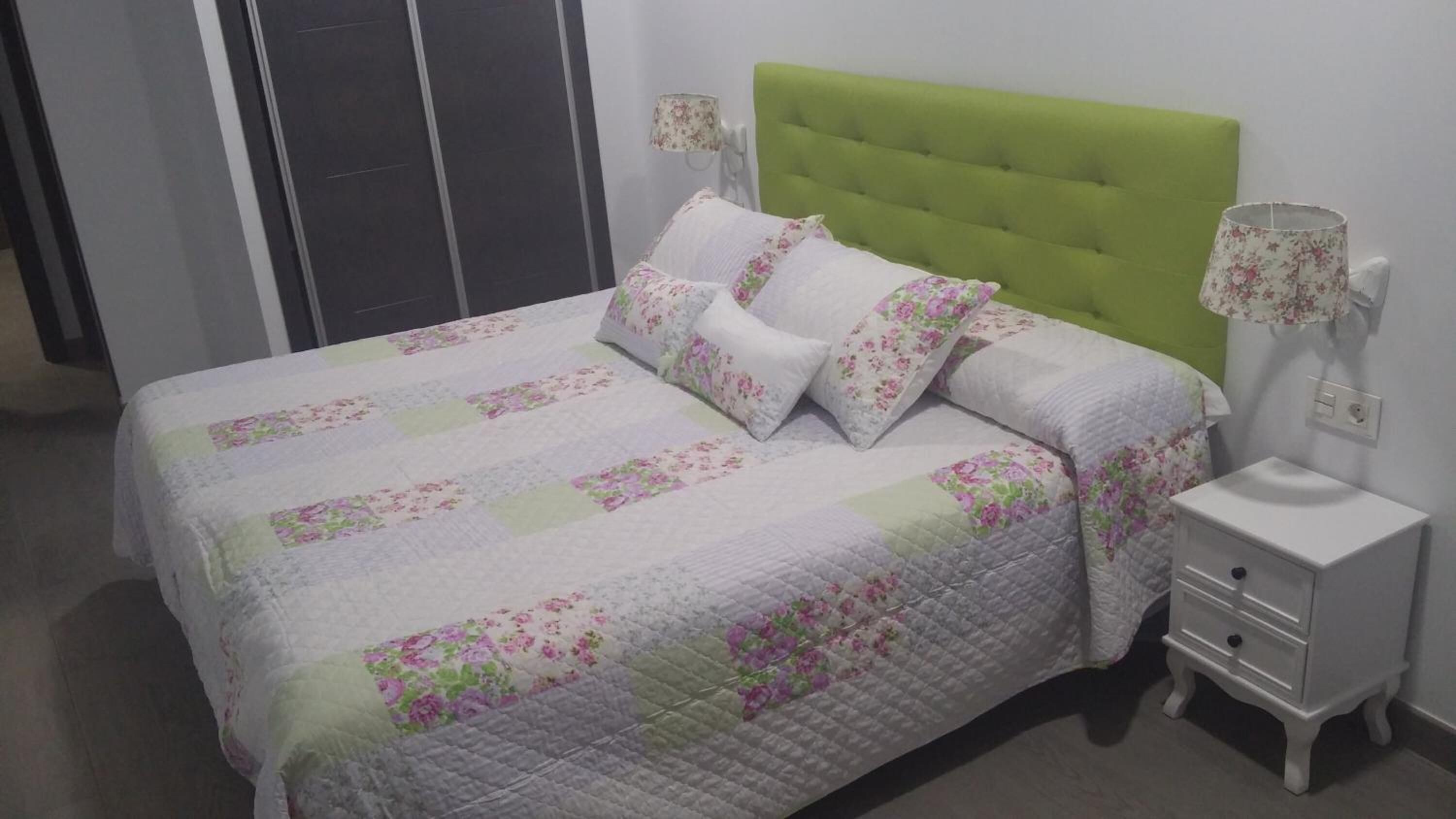 Una cama bien hecha con ropa de cama floral, un cabecero tapizado en verde y una mesita de noche con una lámpara.