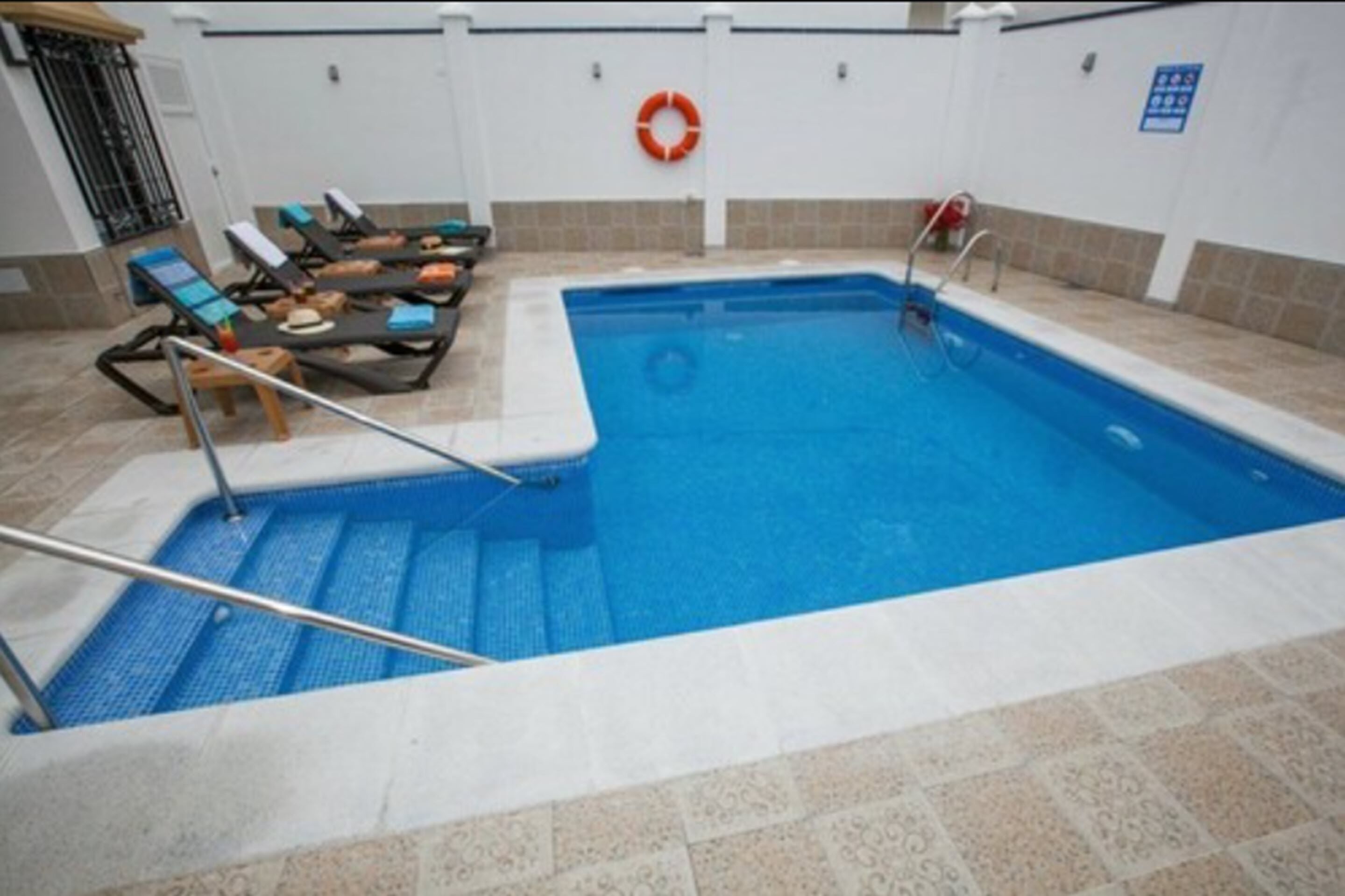 An indoor swimming pool with steps leading into the water, lounge chairs, and a lifebuoy on the wall.