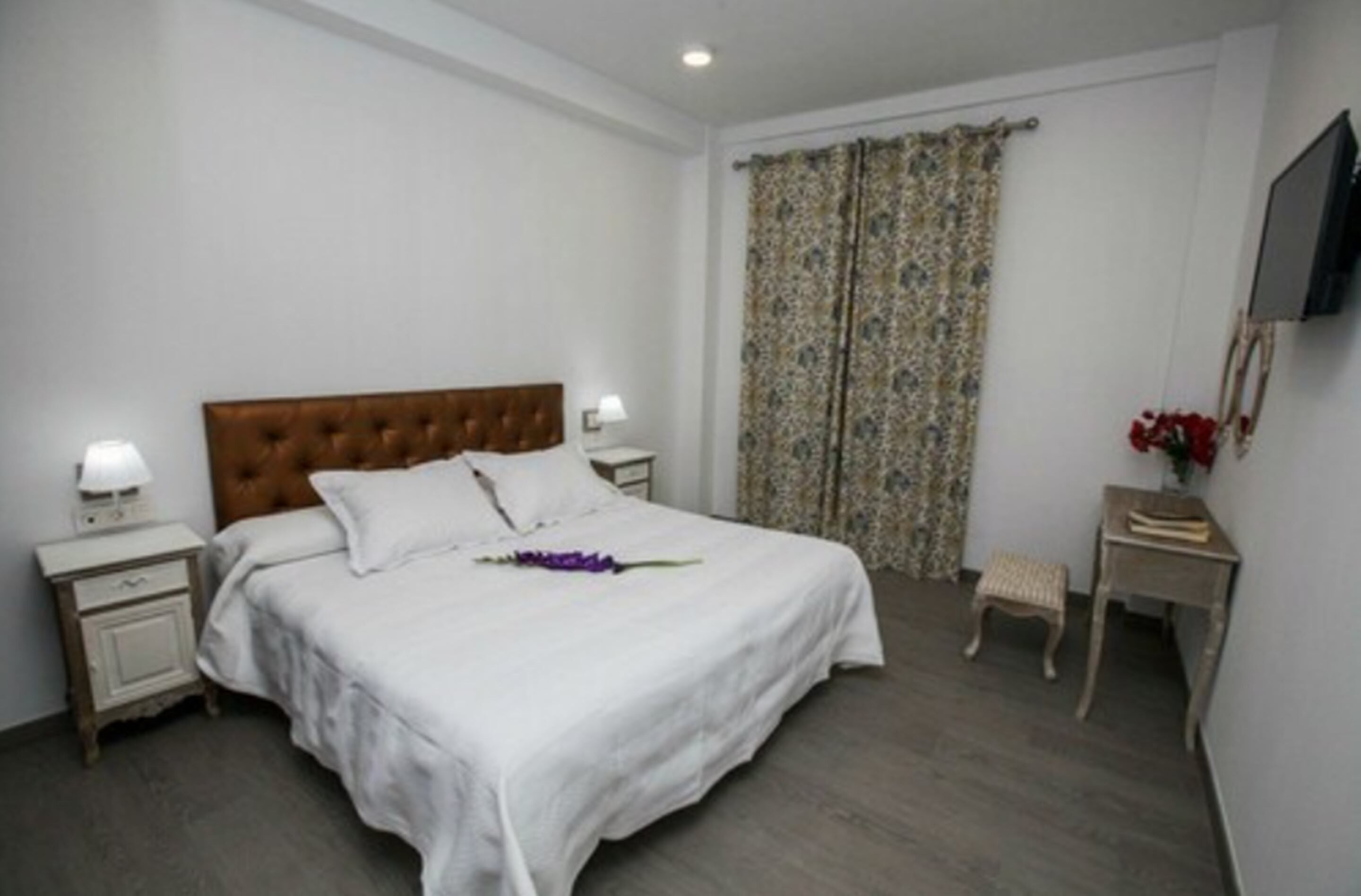 A bedroom with a large bed, two bedside tables, a nightstand, a small stool, and a television on the wall.