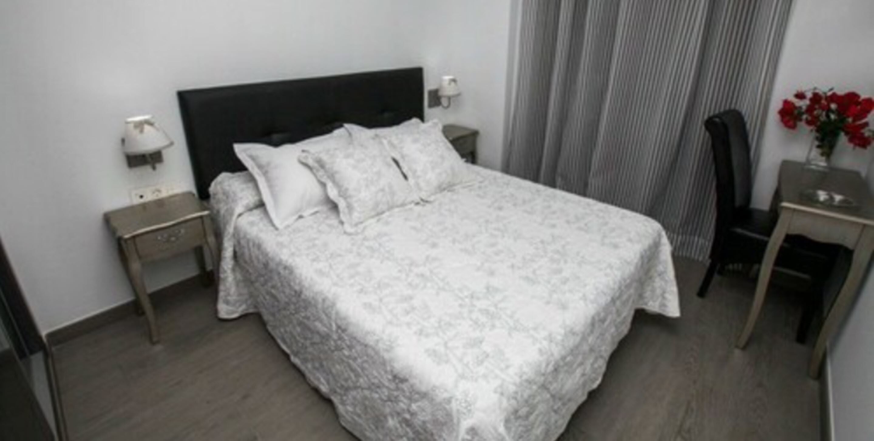 Un lit bien fait, recouvert d’une couvre-lit à motifs, une table de chevet avec une lampe et une chaise dans une pièce aux murs unis et aux rideaux simples.