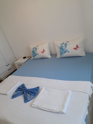 A bed with a blue mattress, two pillows with floral designs and butterflies, a white blanket, and a folded blue towel. - Beyaz Apart-Pansiyon (Cesme)