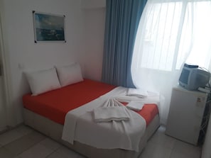 A single bed with a red mattress and white bedding, a small bedside table with a television, a painting on the wall, and a window with curtains. - Beyaz Apart-Pansiyon (Cesme)