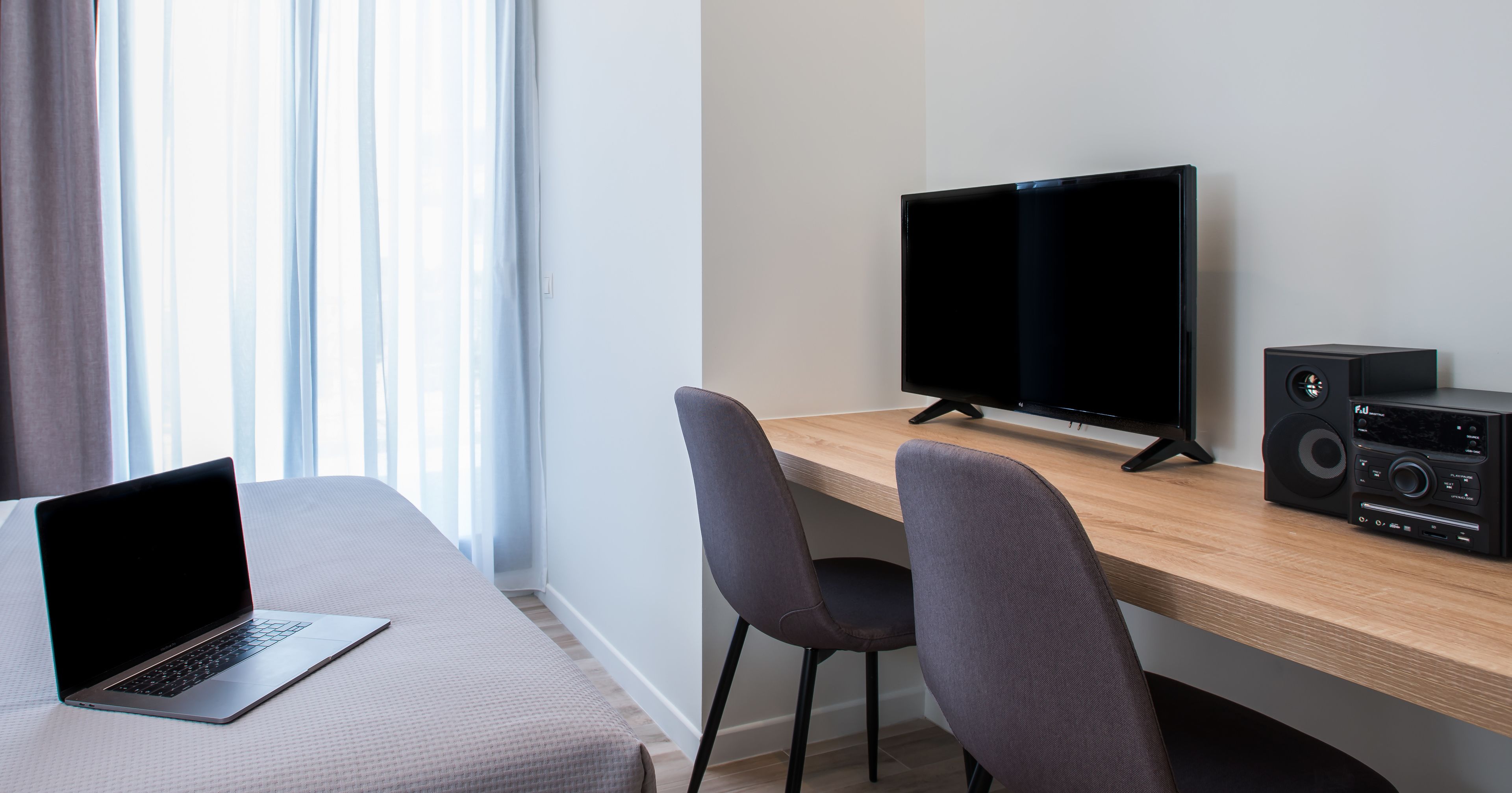 A modern hotel room with a bed, a desk with a laptop, a TV, and a stereo system.