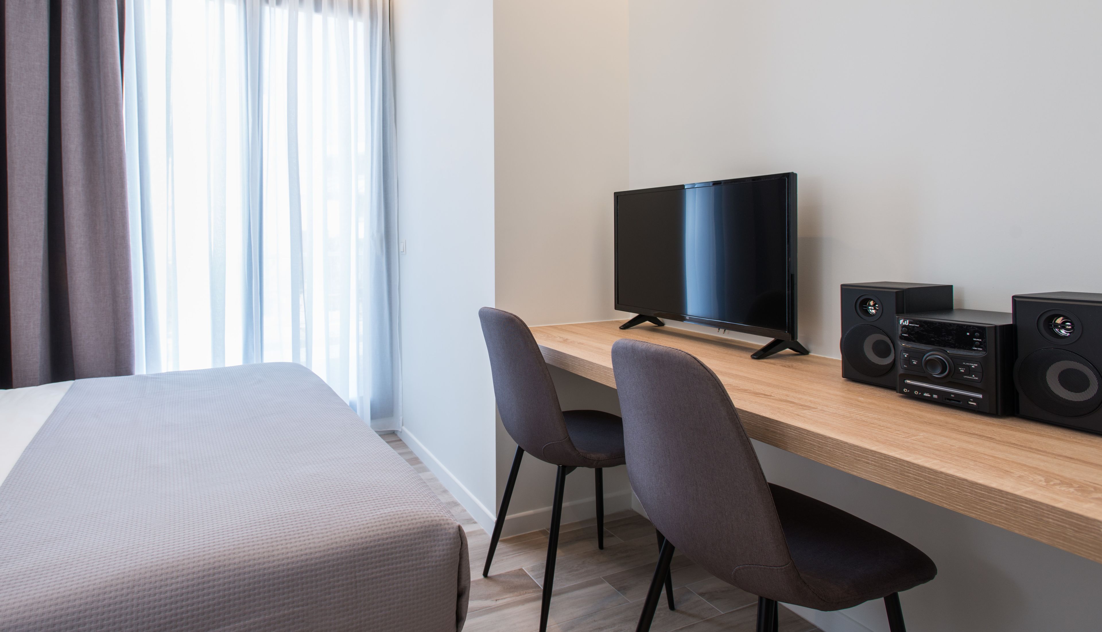A hotel room with a bed, a desk with a computer and speakers, and a window with curtains.