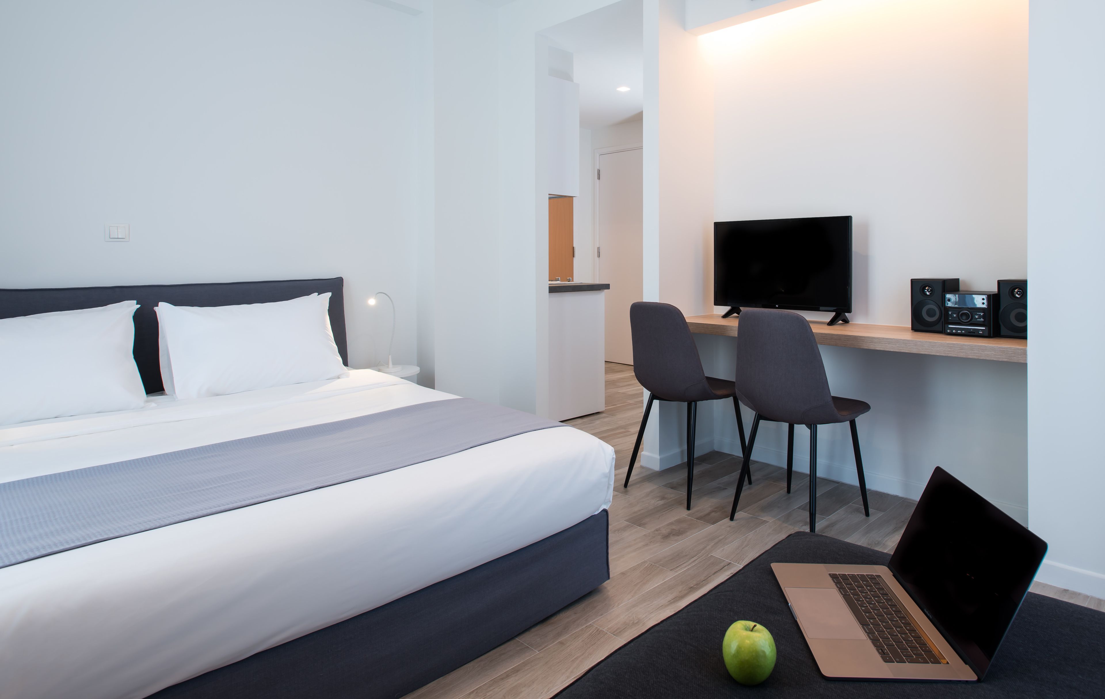 A modern hotel room with a bed, a desk with a laptop, a TV, and speakers.