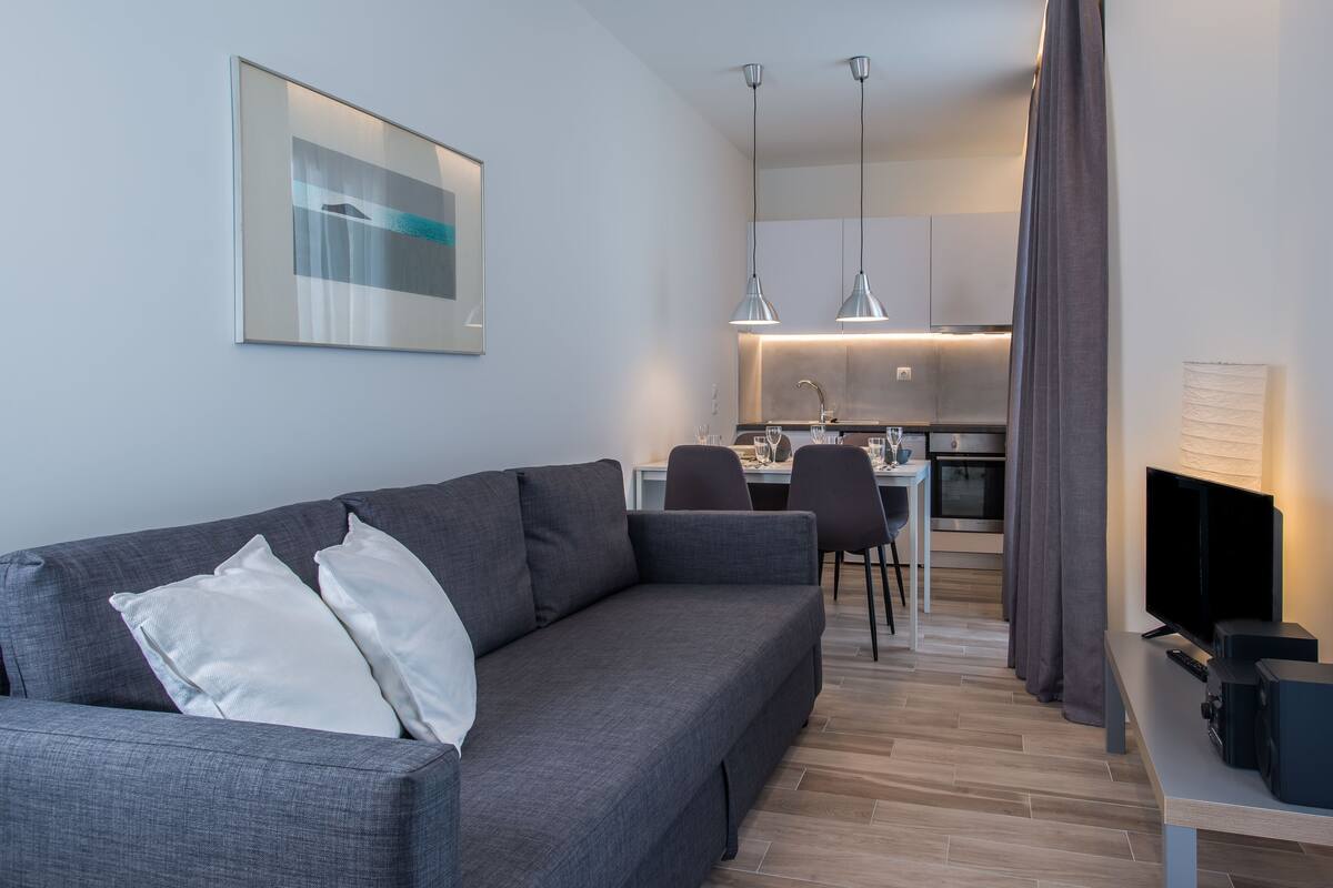A modern living room with a grey sofa, a dining area with a table and chairs, a kitchen with pendant lights, and a flat-screen TV.